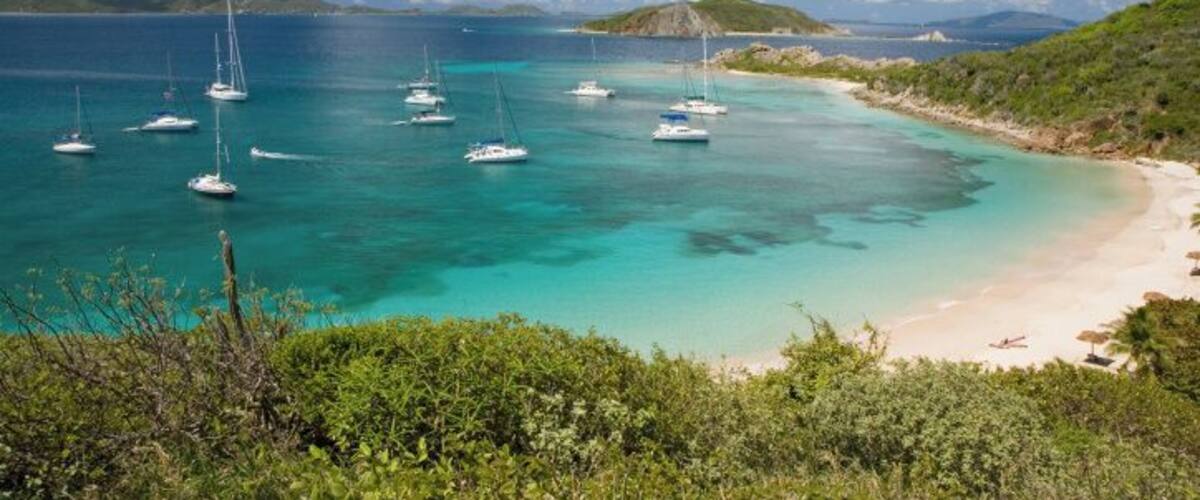 A turquoise sea with sailboats, a sandy beach, and green hills.