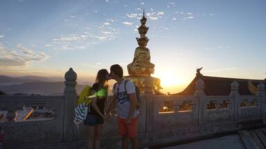 Um casal se beijando em frente a uma grande estátua dourada ao pôr do sol.