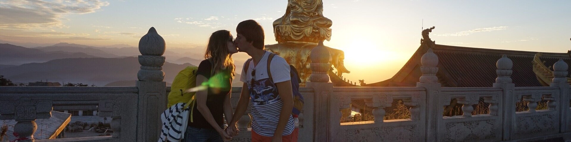 Um casal se beijando em frente a uma grande estátua dourada ao pôr do sol.