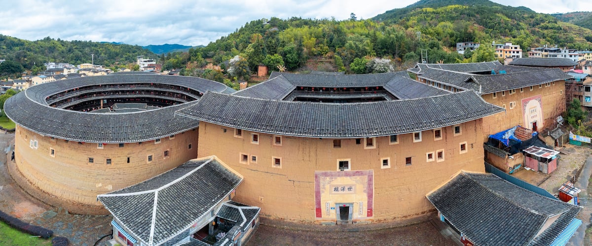 A traditional round building with multiple levels and a tiled roof, surrounded by a hilly landscape and other buildings.