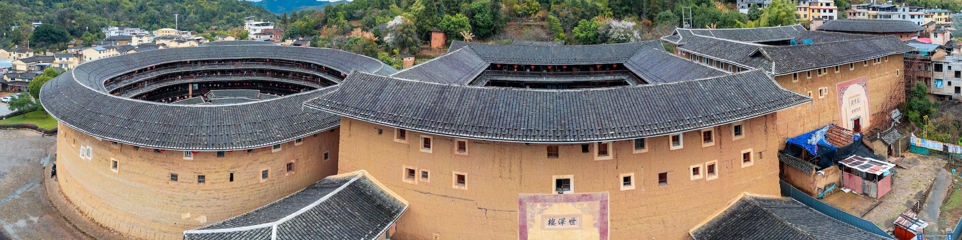 A traditional round building with multiple levels and a tiled roof, surrounded by a hilly landscape and other buildings.