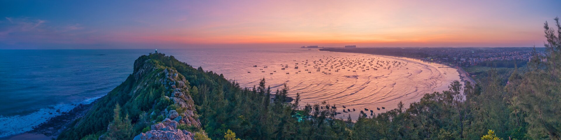 A coastal landscape with a rocky outcrop, dense forest, and a beach with numerous boats.