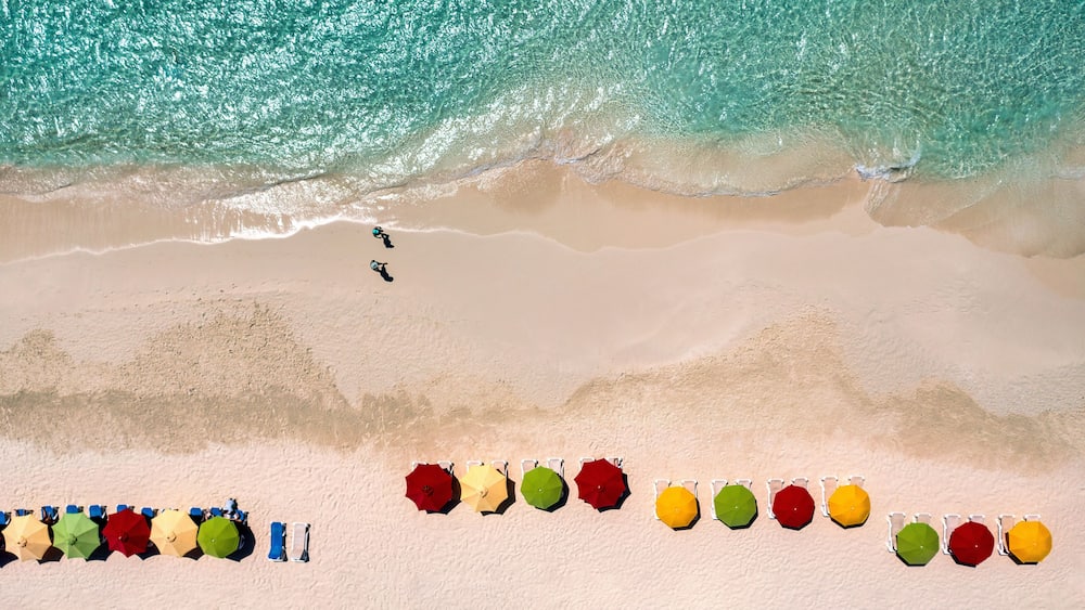Une vue aérienne d’une plage aux eaux turquoise, avec un rivage sablonneux et des parasols colorés alignés le long de la plage.