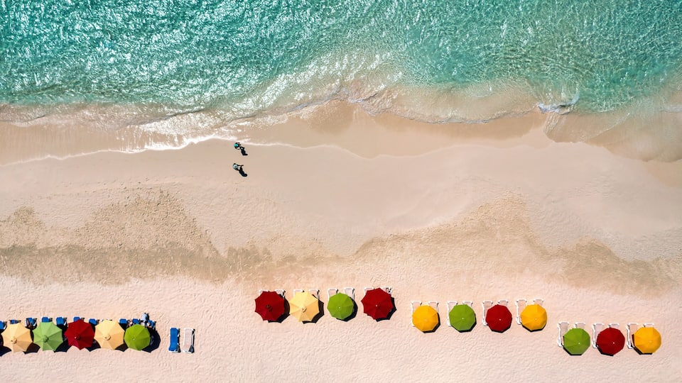 An aerial view of a beach with turquoise water, a sandy shore, and colorful umbrellas lined up along the beach.