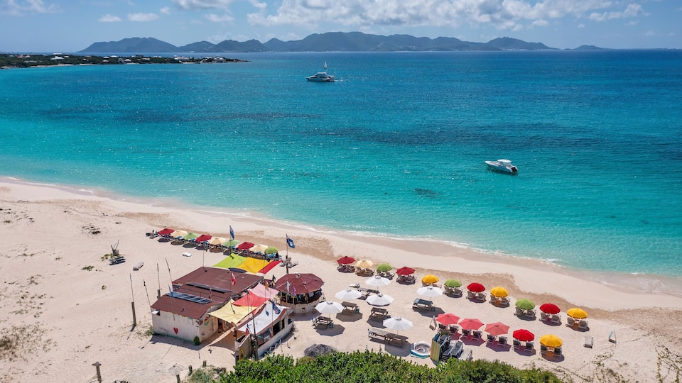A beach with clear turquoise waters, a boat in the distance, and colorful umbrellas and beach chairs.