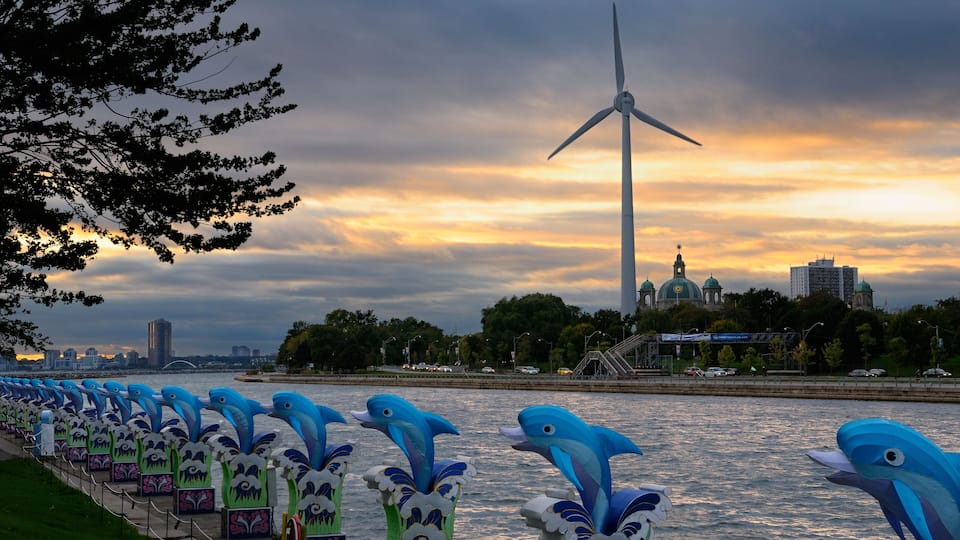 A river with a wind turbine and a row of dolphin sculptures along the riverbank.