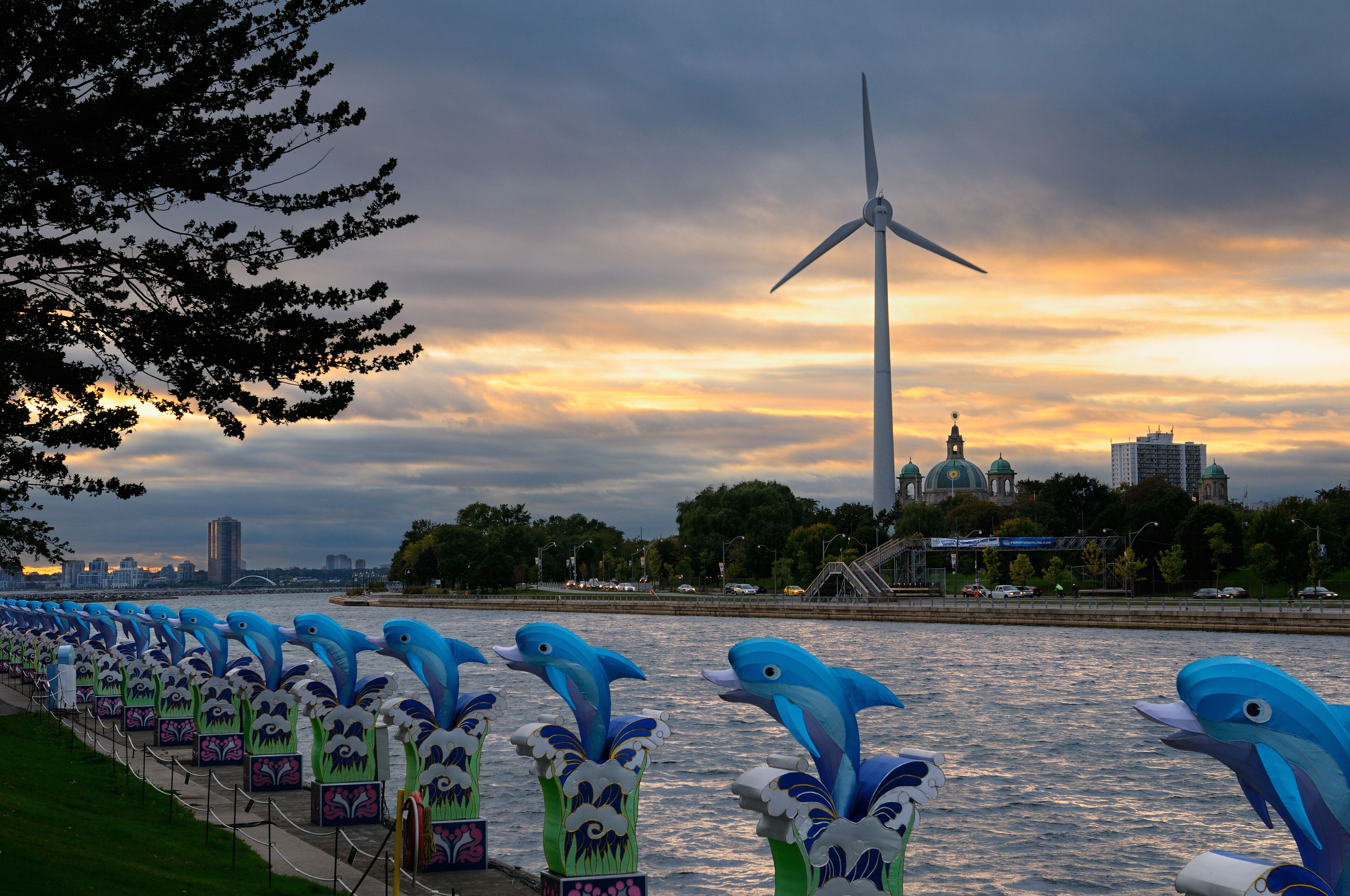 A river with a wind turbine and a row of dolphin sculptures along the riverbank.