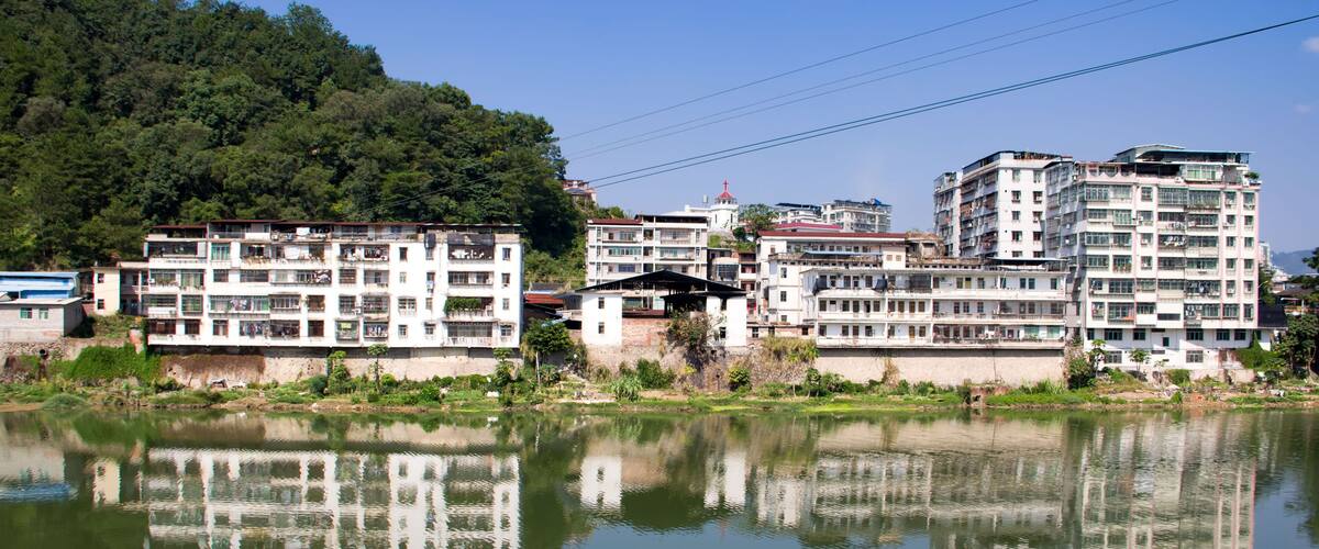 A calm river reflecting buildings and a forested hill.