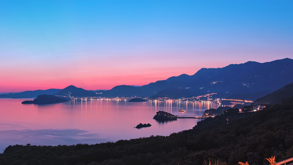 A coastal landscape at dusk with a calm sea, illuminated city lights, and distant mountains.