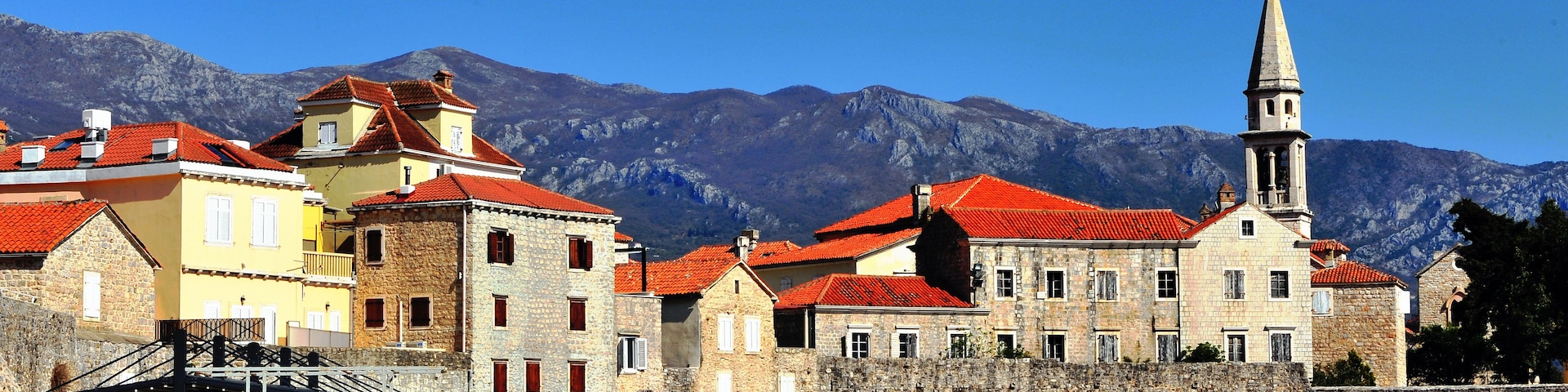 Uma vila costeira com edifícios de pedra, uma igreja com uma alta torre sineira e montanhas ao fundo.
