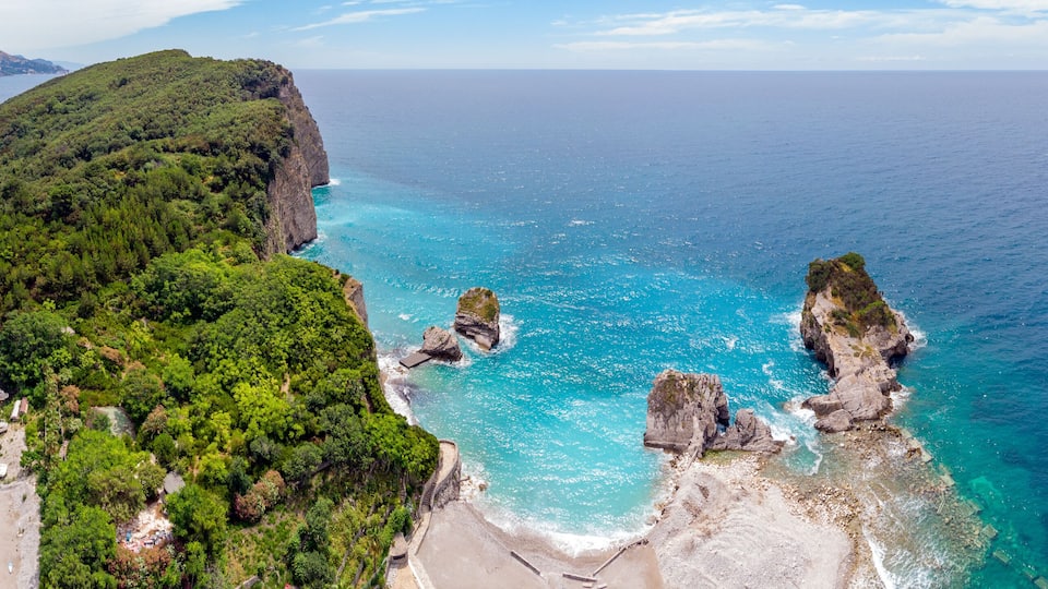 A coastal landscape with a rocky bay, a forested hill, and a marina with boats.