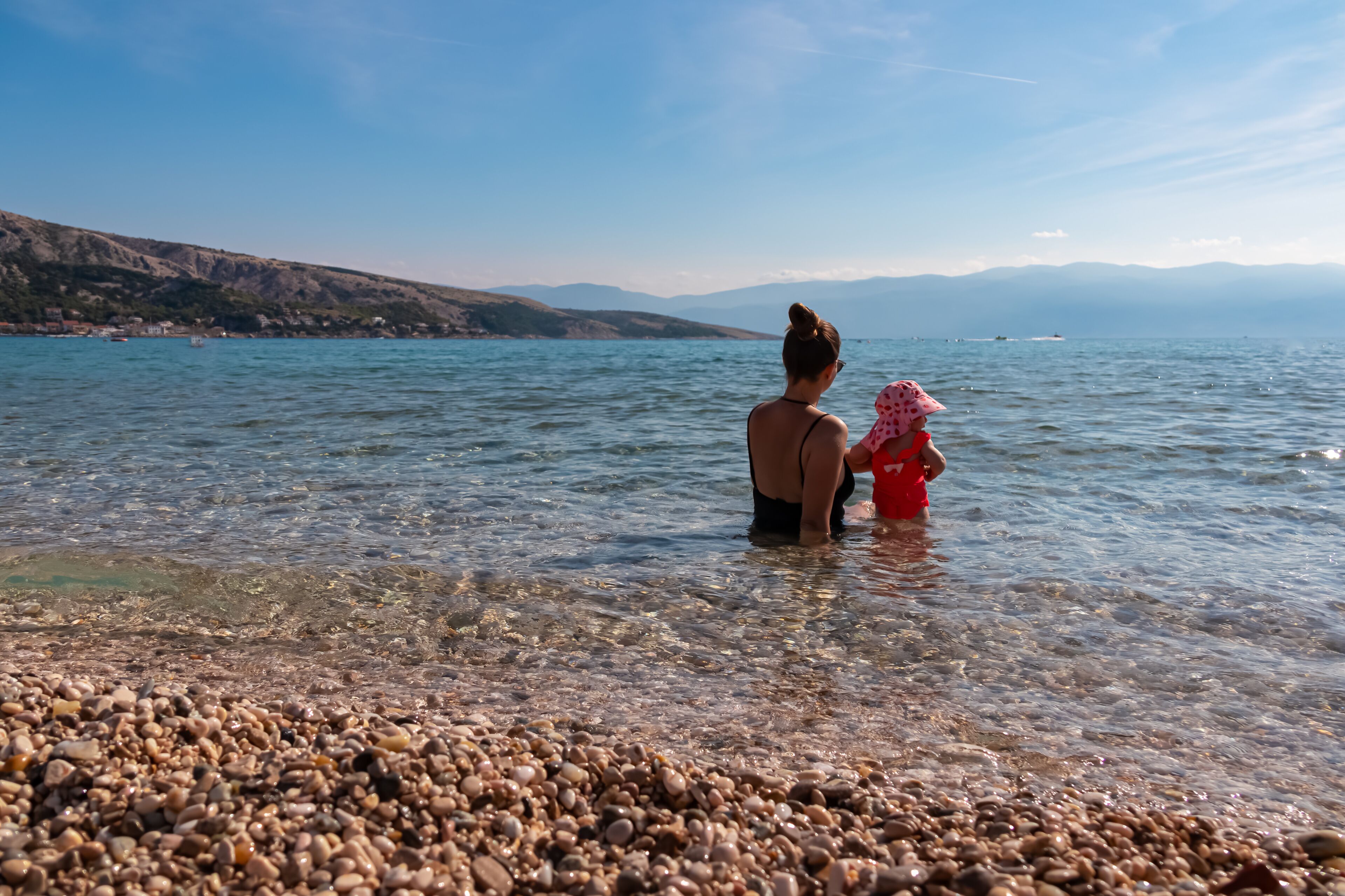 Loving mother swimming with toddler on paradise pebble beach in coastal town Baska, Krk Island, Primorje-Gorski Kotar, Croatia, Europe. Scenic view of coastline of Mediterranean Adriatic Sea in summer