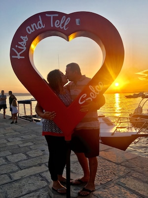 A couple embracing in front of a heart-shaped sign with the words "Kiss and Tell" against a sunset backdrop.