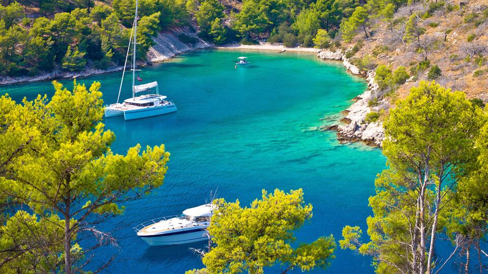 A turquoise bay with boats anchored near the shore, surrounded by dense pine trees and rocky cliffs.