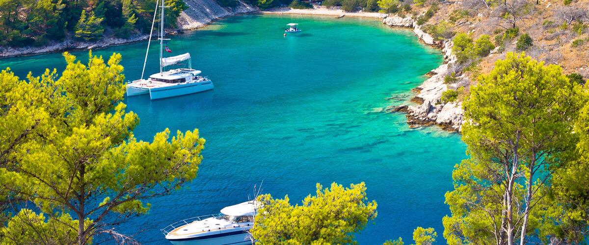 A turquoise bay with boats anchored near the shore, surrounded by dense pine trees and rocky cliffs.