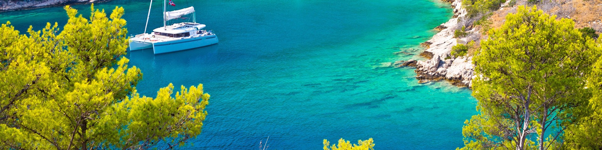 A turquoise bay with boats anchored near the shore, surrounded by dense pine trees and rocky cliffs.