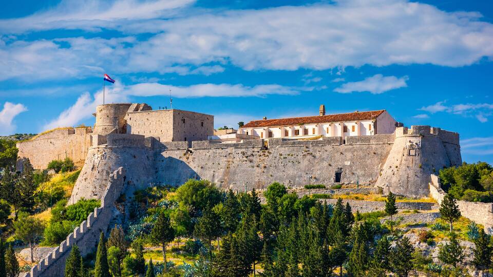A historic stone fortress with a flag, surrounded by lush greenery and a clear blue sky.