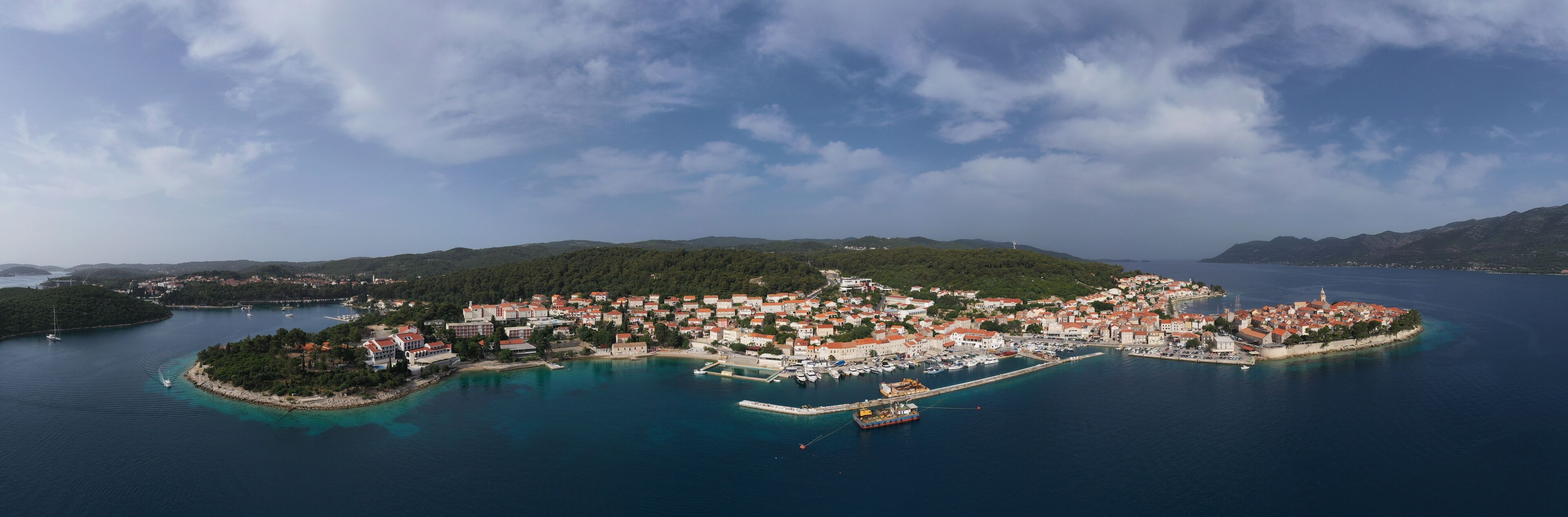An aerial view of a coastal town with a marina, surrounded by hills and clear blue waters.