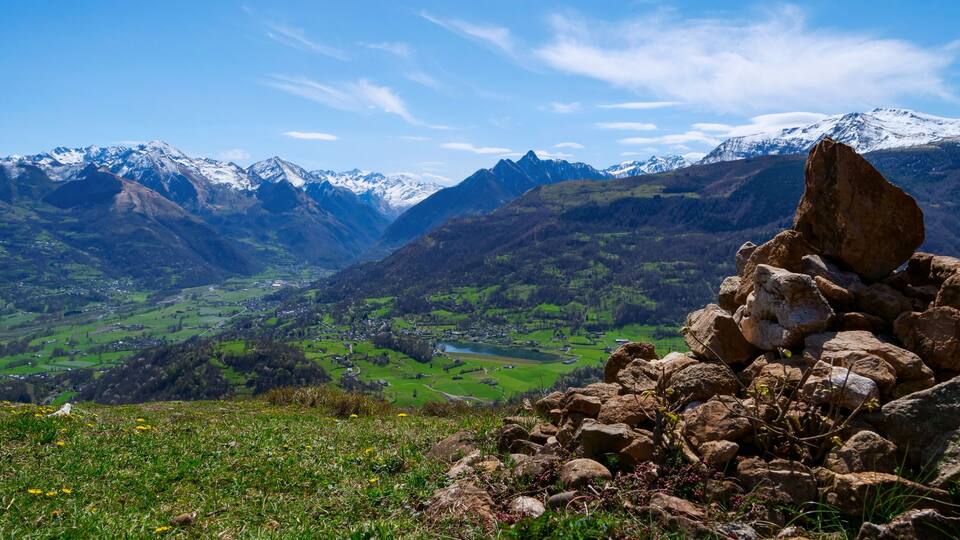 A mountainous landscape with snow-capped peaks, a valley with green fields, and a rocky outcrop in the foreground.