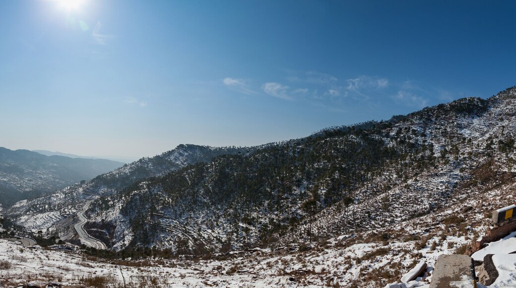 View of snow-dusted mountains and winding roads under a bright sun, a serene winter landscape in Murree, Punjab, Pakistan.