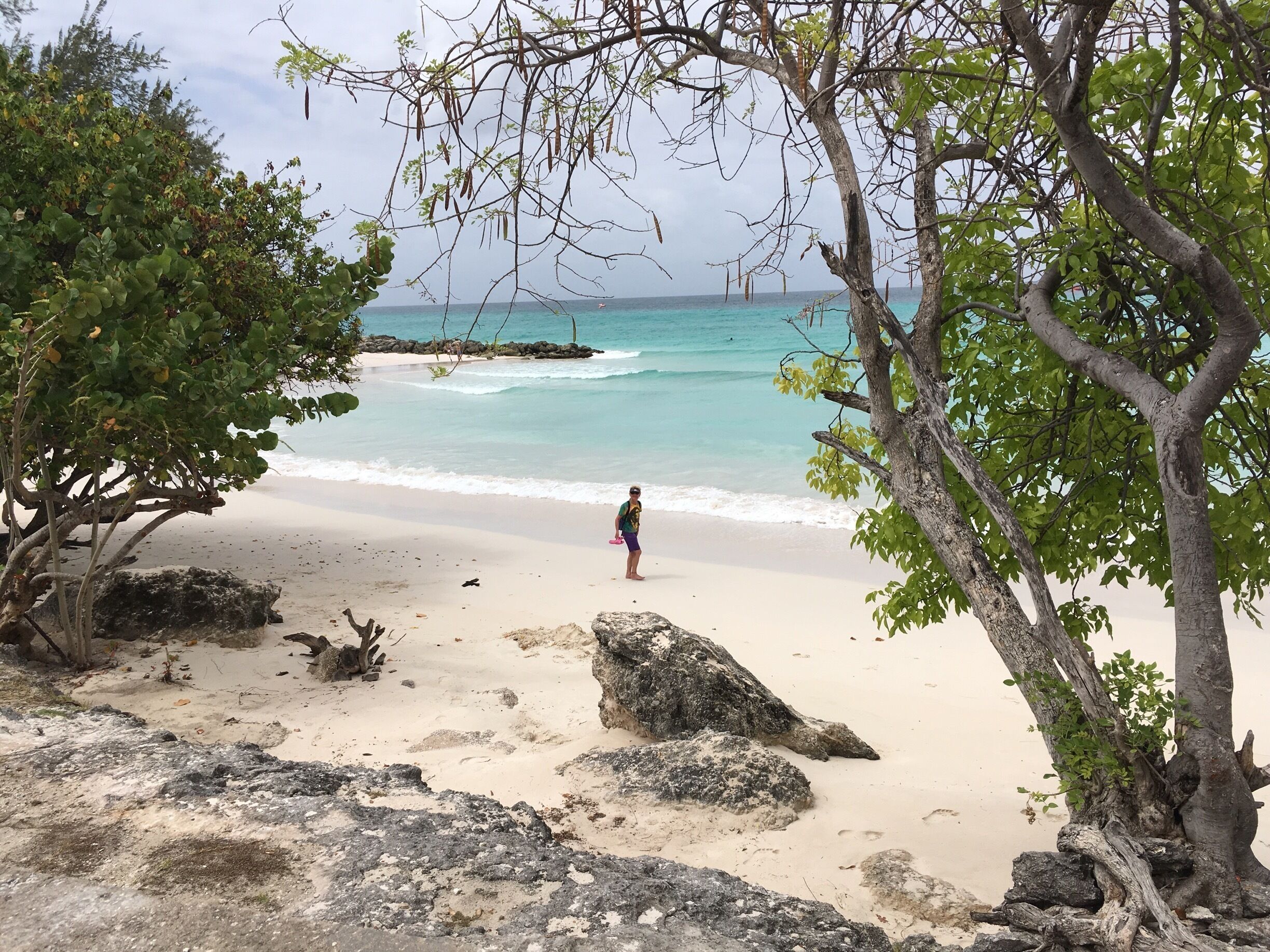 A beach with clear turquoise water, a person standing on the shore, and rocky outcrops.