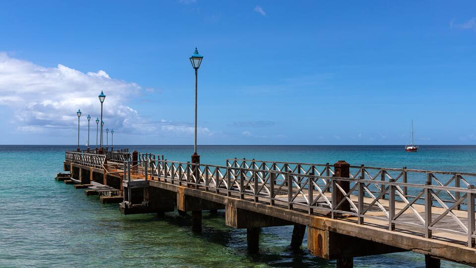 A wooden pier extending into the ocean with clear water, a sailboat in the distance, and a clear blue sky.