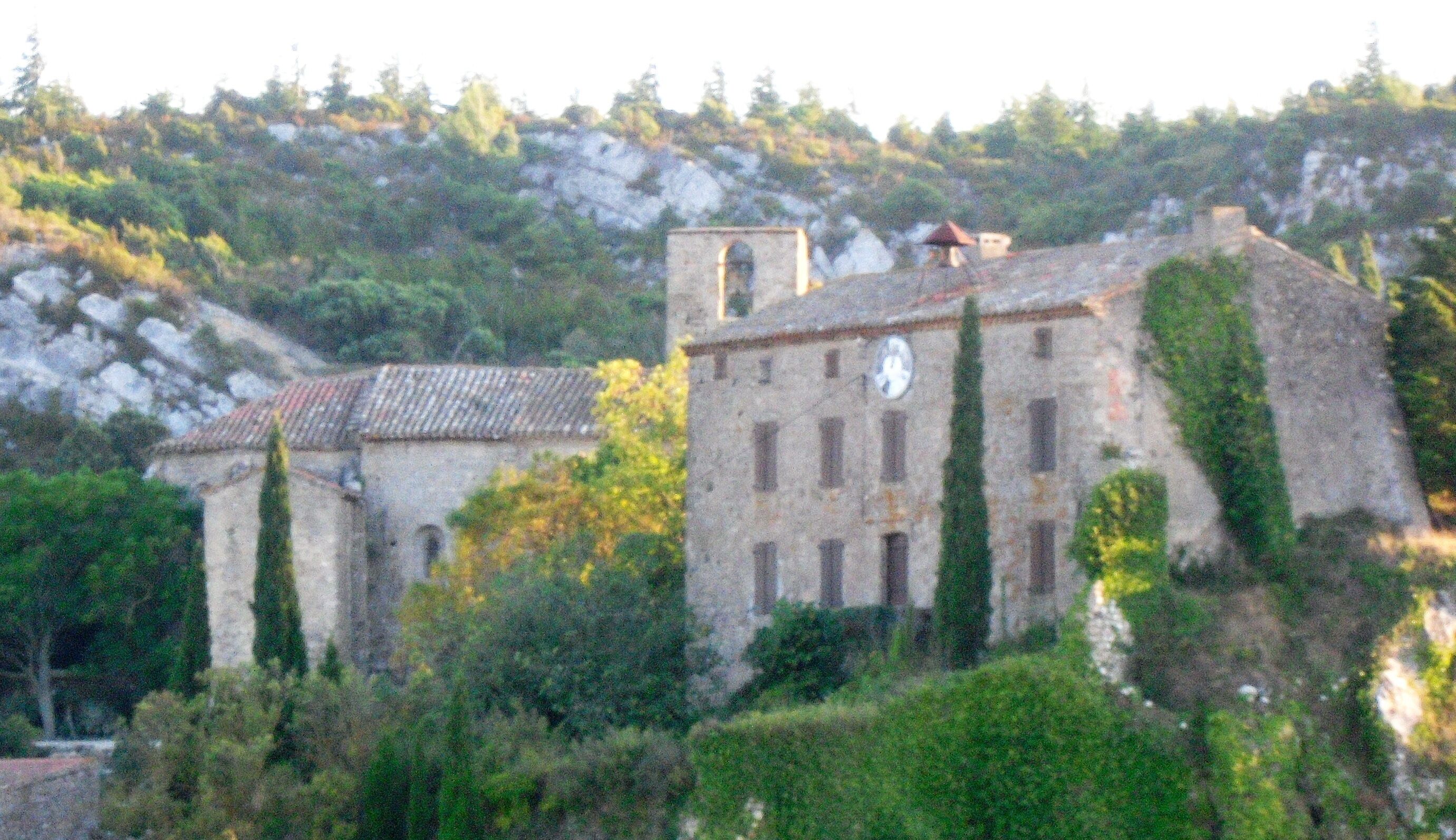 A historic stone building with a clock tower, surrounded by lush greenery and rocky terrain.