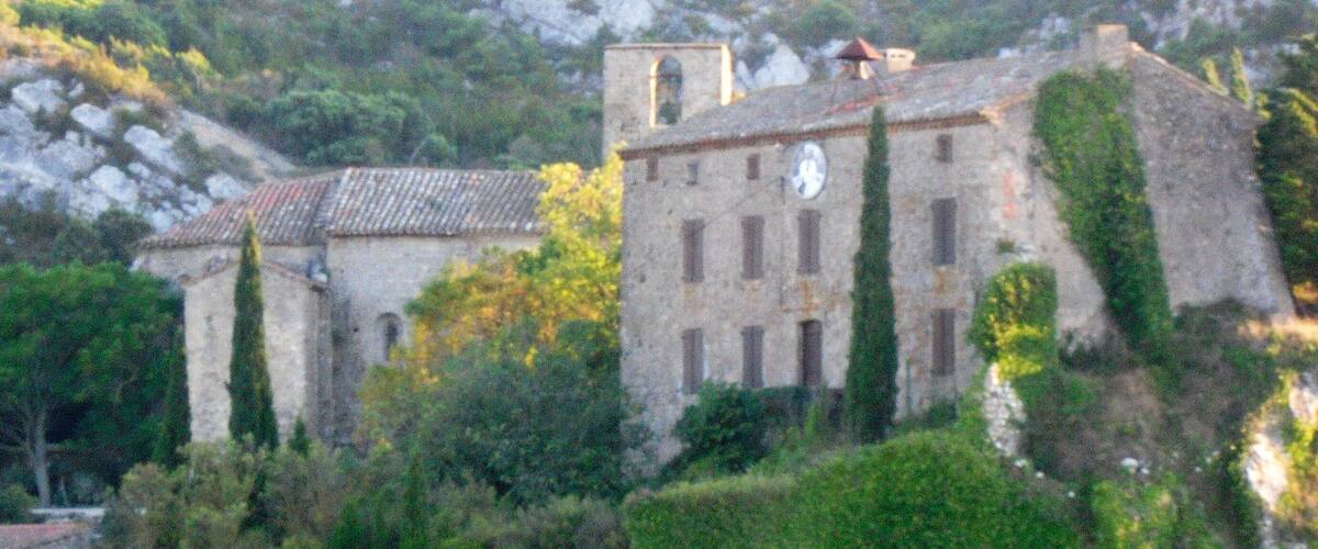 A historic stone building with a clock tower, surrounded by lush greenery and rocky terrain.