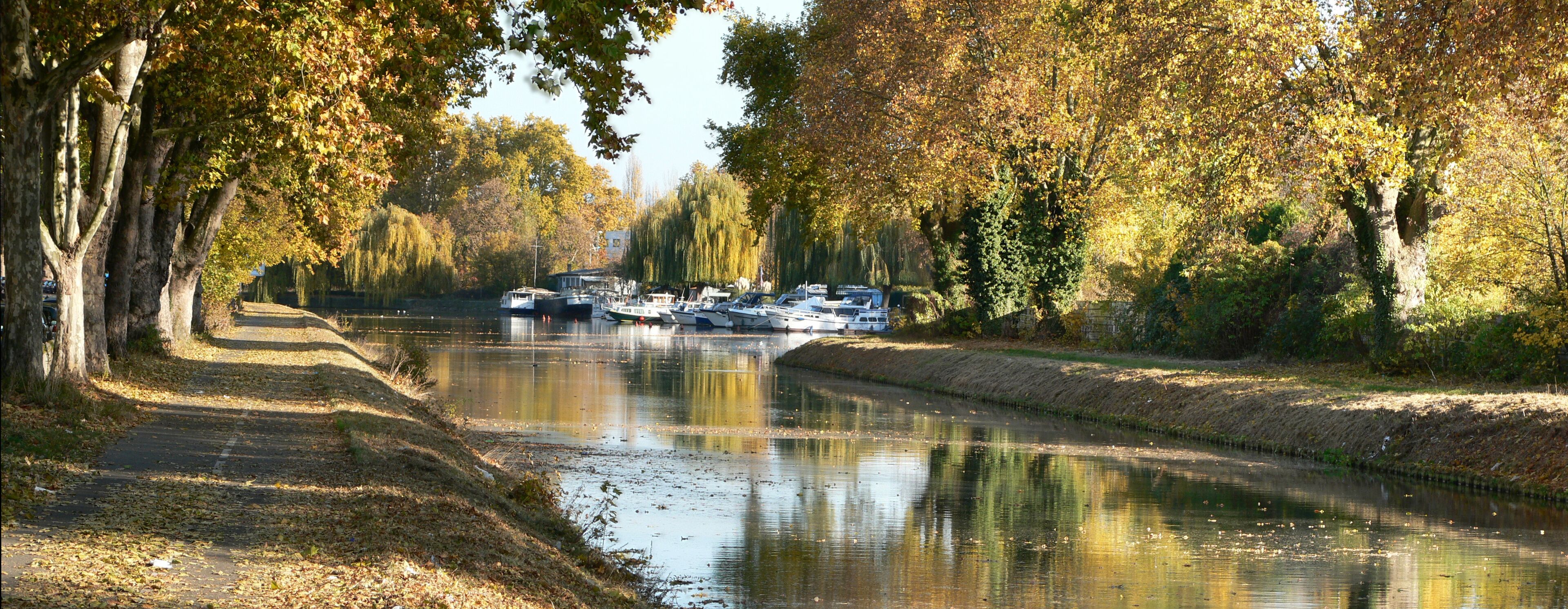 A canal lined with trees reflecting boats and a path.