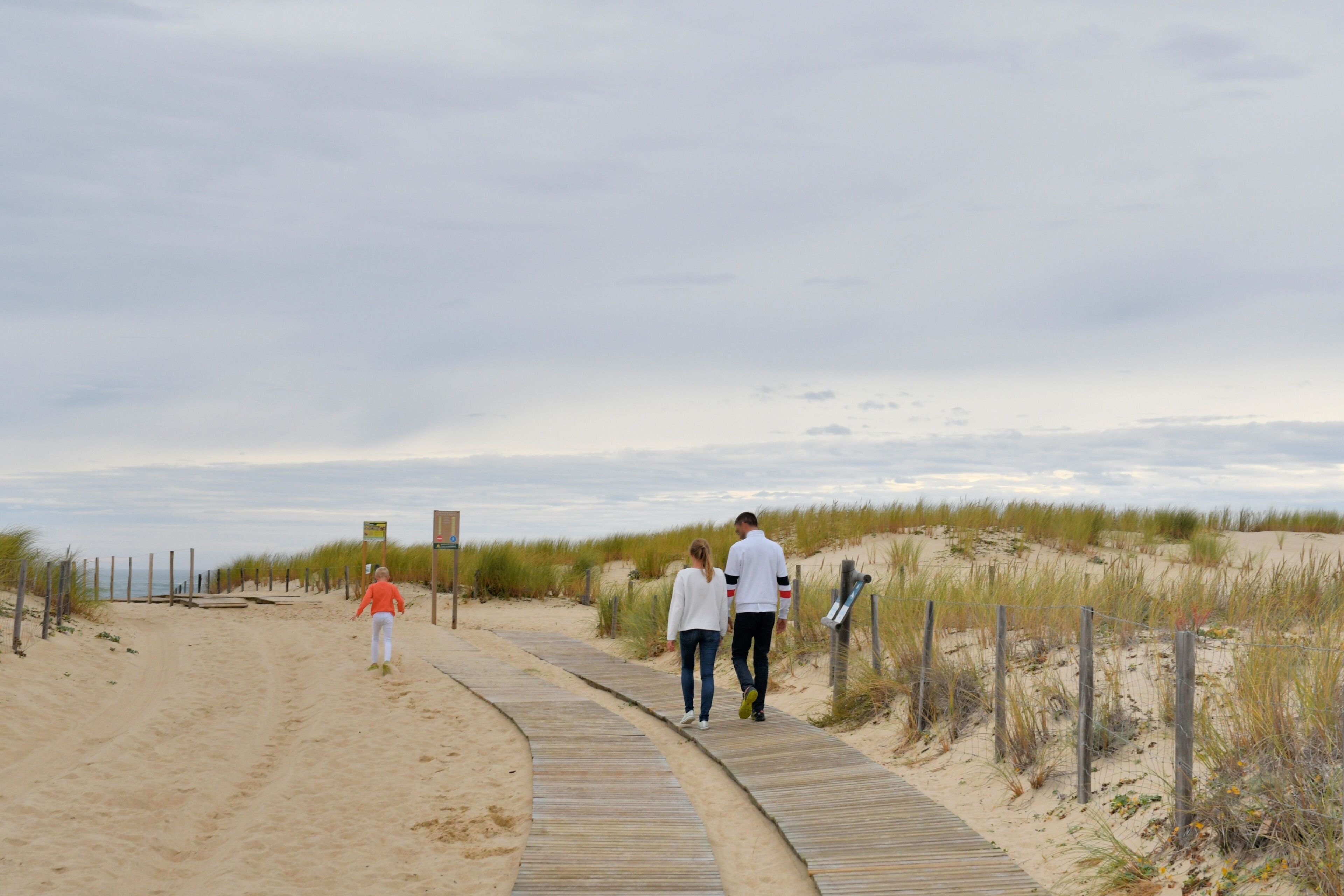 A sandy beach path with people walking, surrounded by dunes and a cloudy sky.