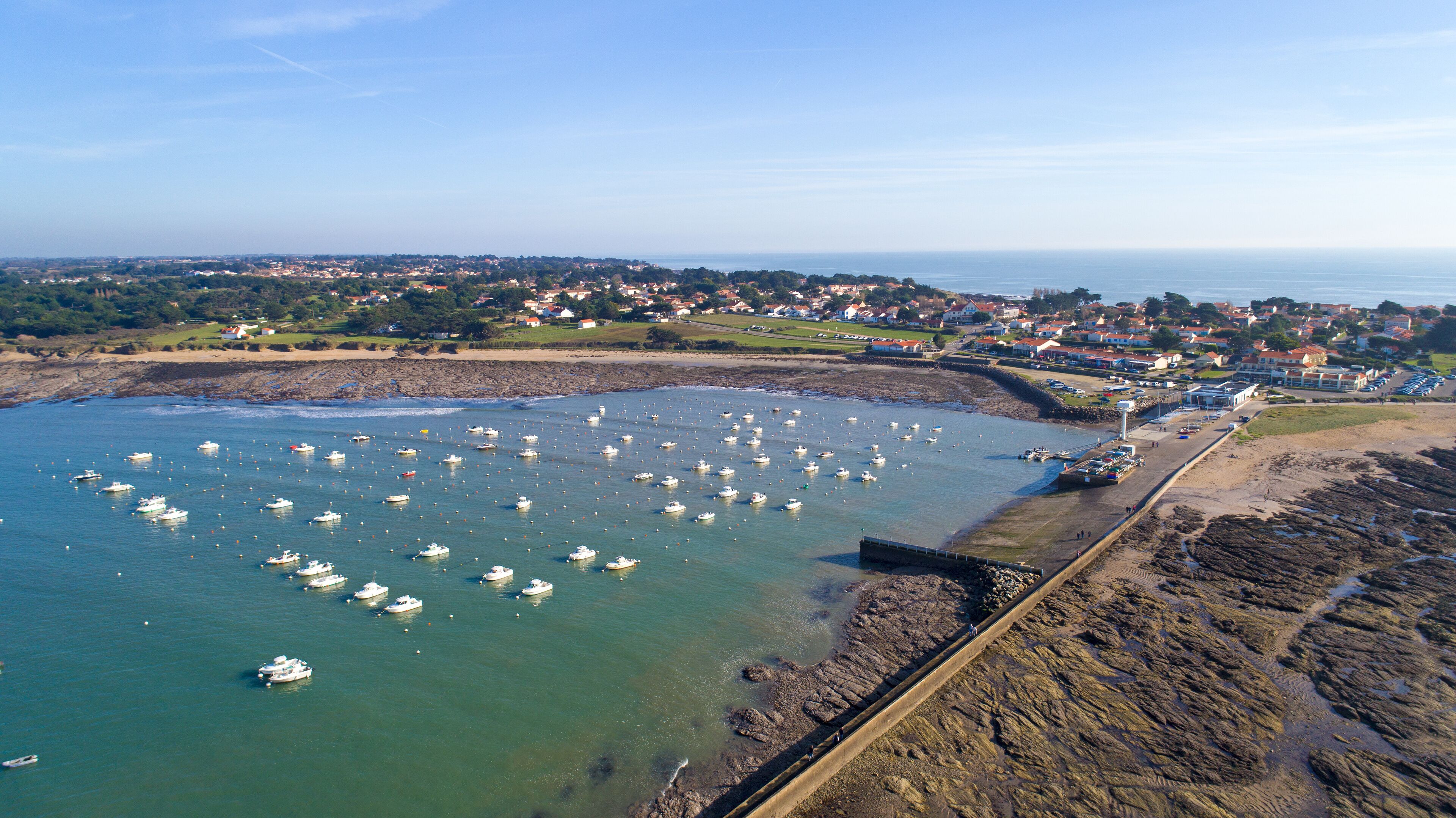 A coastal town with a harbor filled with boats, a rocky shoreline, and residential buildings in the background.