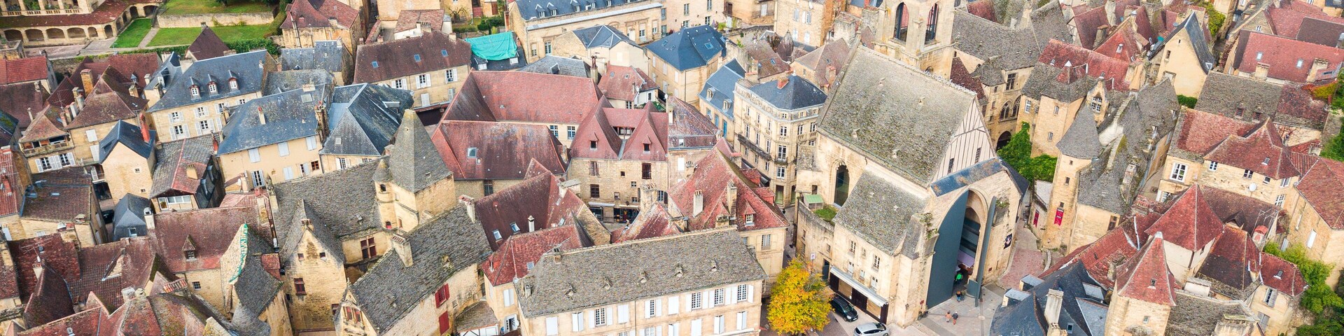 Vista aérea de uma cidade histórica europeia, com edifícios bem próximos, ruas de paralelepípedos e uma igreja imponente com um grande arco.