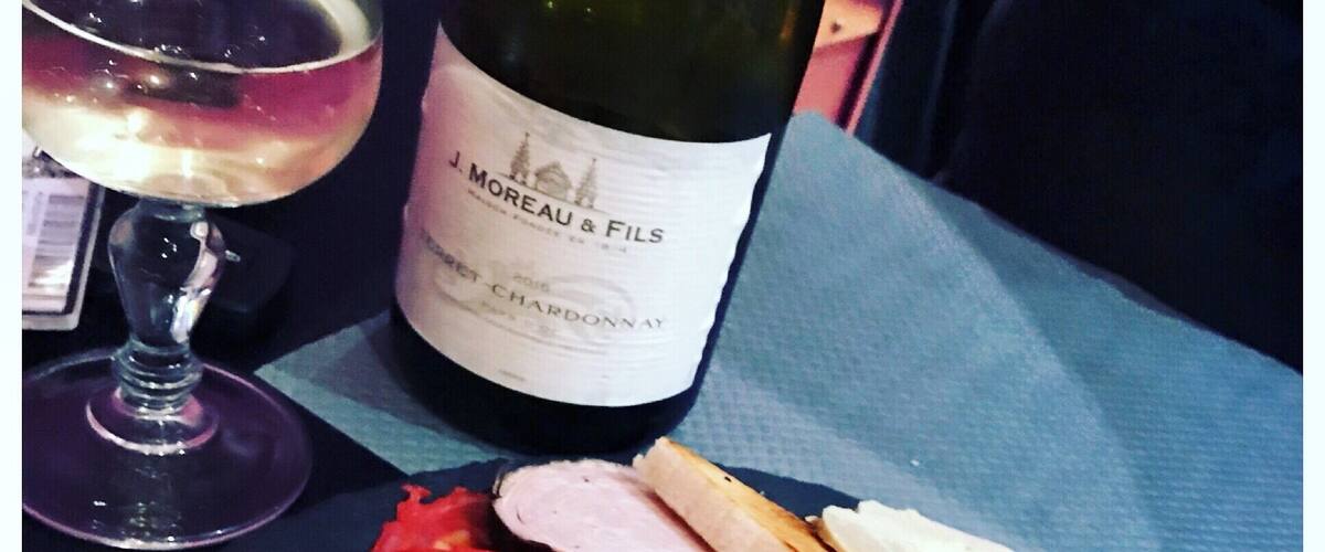 A bottle of J. Moreau & Fils 2016 Pet-Chardonnay, a glass of wine, and a plate with various meats and pickles.