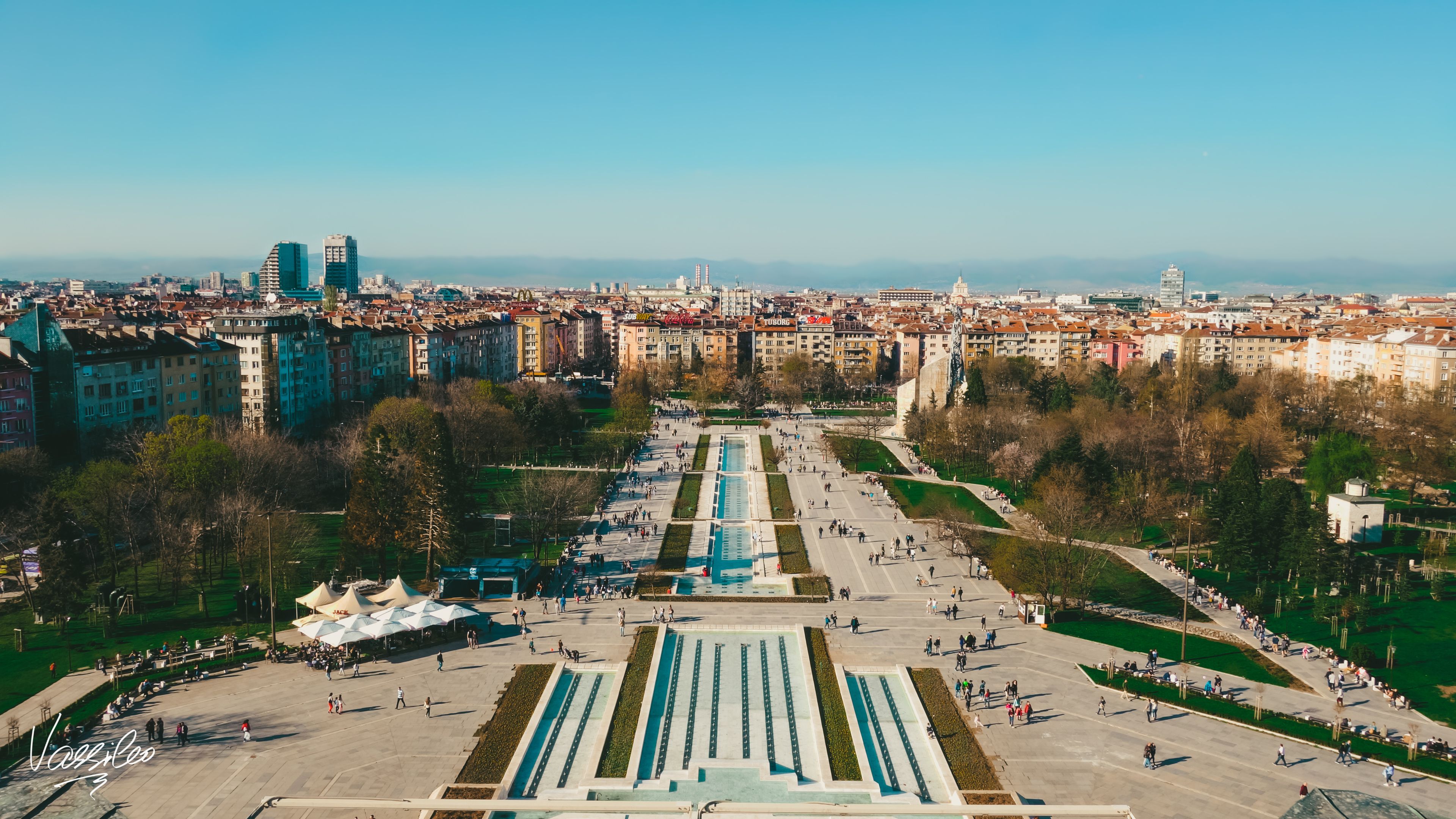 Paisagem urbana com uma praça central, edifícios ao redor e uma cadeia de montanhas ao longe.