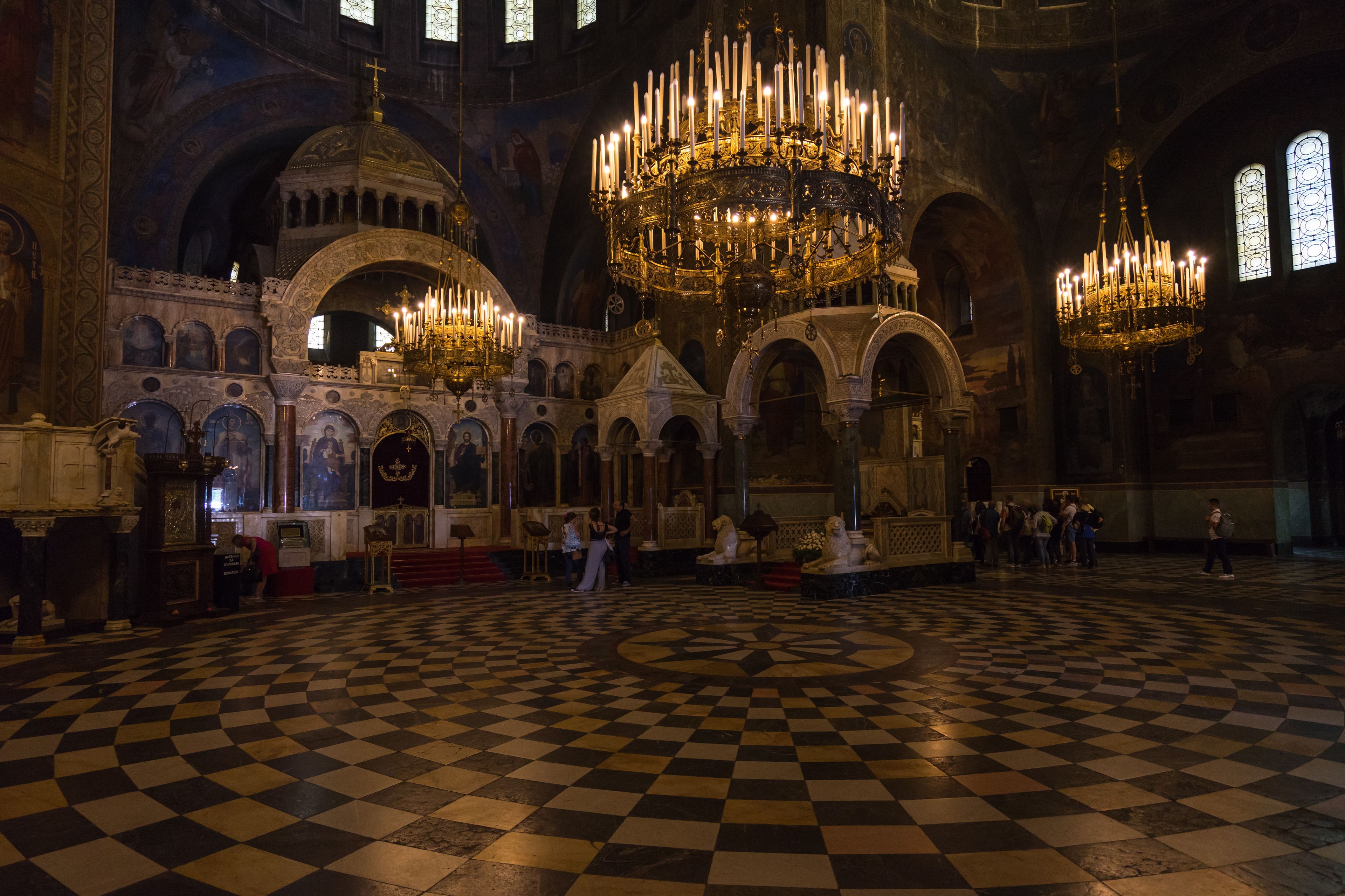 Interior de igreja ornamentado, com altos tetos, lustres e chão xadrez.