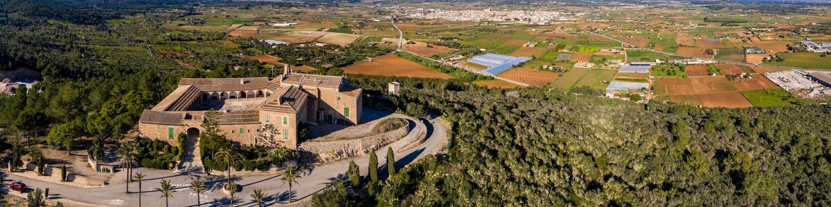 A historic building with a curved pathway, surrounded by dense forest and agricultural fields.