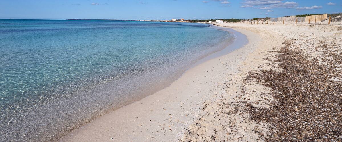 A clear blue sea, sandy beach, and a few scattered buildings in the distance.
