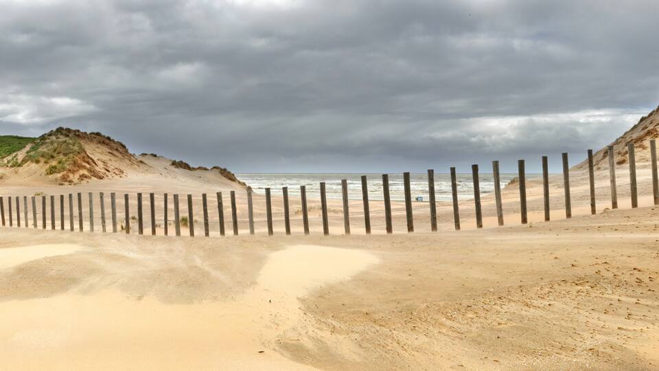 A sandy beach with a wooden fence and dunes under a cloudy sky.