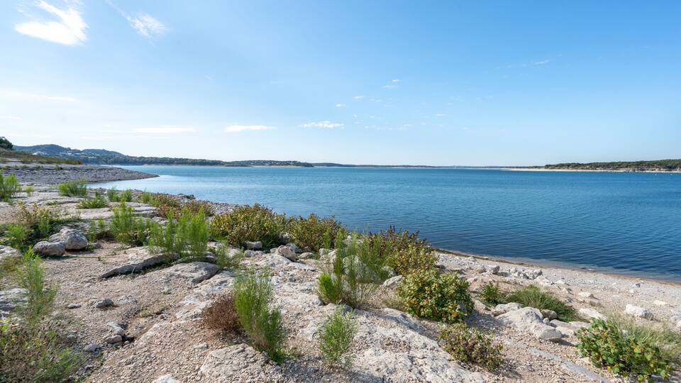 A rocky shoreline with sparse vegetation and calm blue waters extending to a distant landmass under a clear sky.