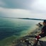 Una persona de pie sobre una costa rocosa, contemplando una masa de agua tranquila con una tierra firme a la distancia.