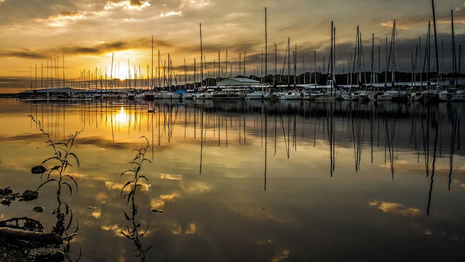 A marina with numerous sailboats docked, reflecting the sunset and clouds in the calm water.