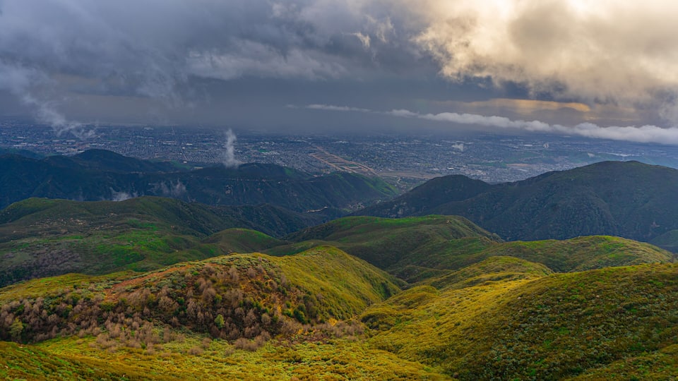 A mountainous landscape with green hills and a city in the distance under a cloudy sky.
