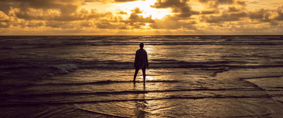 A person standing on a beach facing the ocean during sunset.