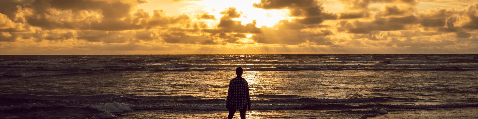 A person standing on a beach facing the ocean during sunset.