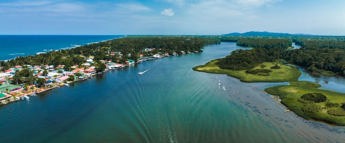 An aerial view of a coastal town with a river, surrounded by dense forests and a clear blue sky.