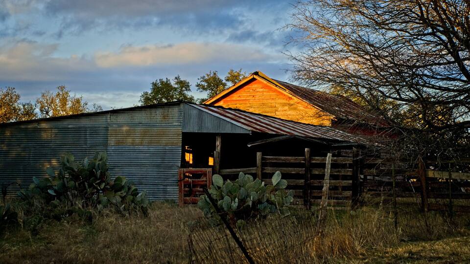 A barn with a wooden roof and corrugated metal walls, surrounded by dry grass and a wooden fence.