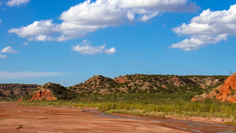 A dry riverbed with red soil, green vegetation, and red rock formations under a clear blue sky.