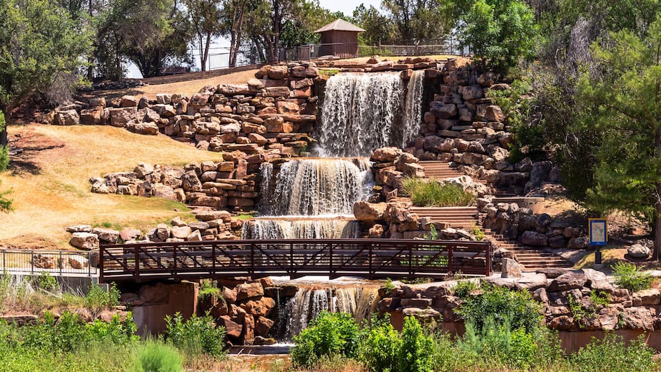 A waterfall cascading over rocks, a wooden bridge, and a small building in a park.
