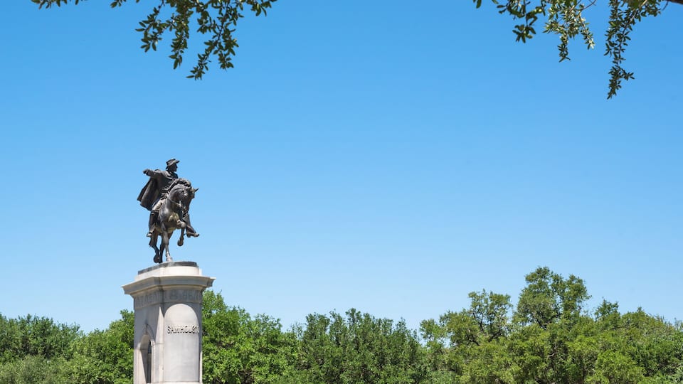 A statue of a mounted figure on a pedestal in a park with green trees and a clear blue sky.