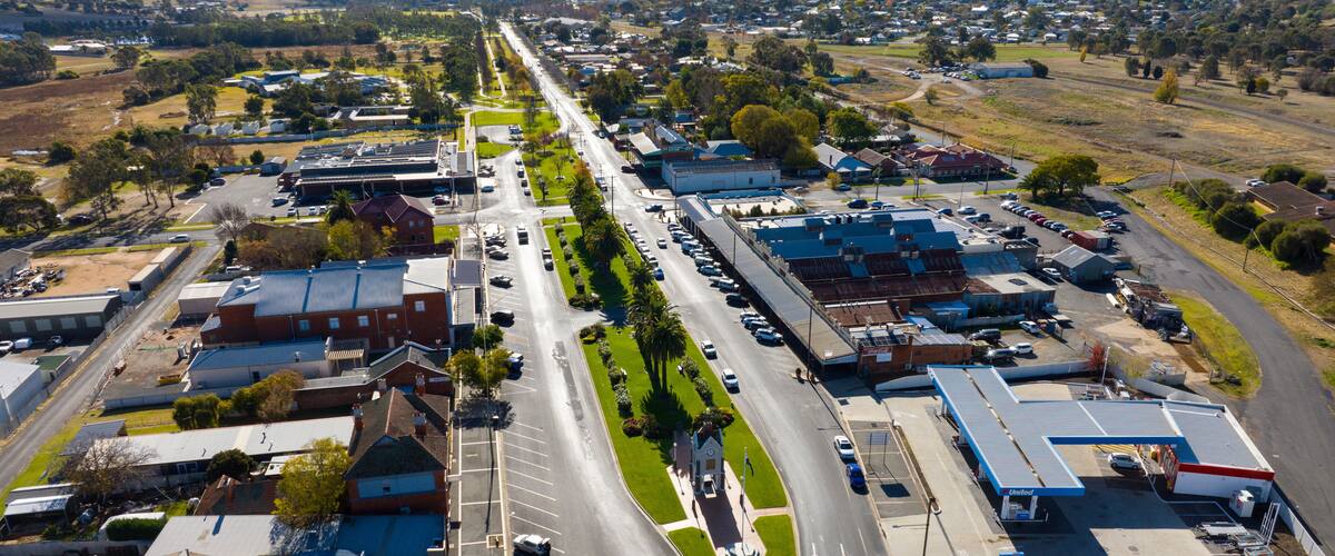 An aerial view of a town with a main road lined with buildings, a parking lot, and surrounding fields.